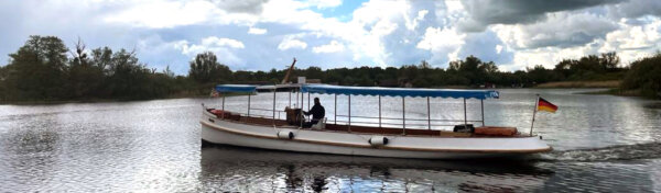 Ein Ausflugsboot mit deutscher Flagge fährt auf ruhigem Gewässer, umgeben von Bäumen und Wolken am Himmel